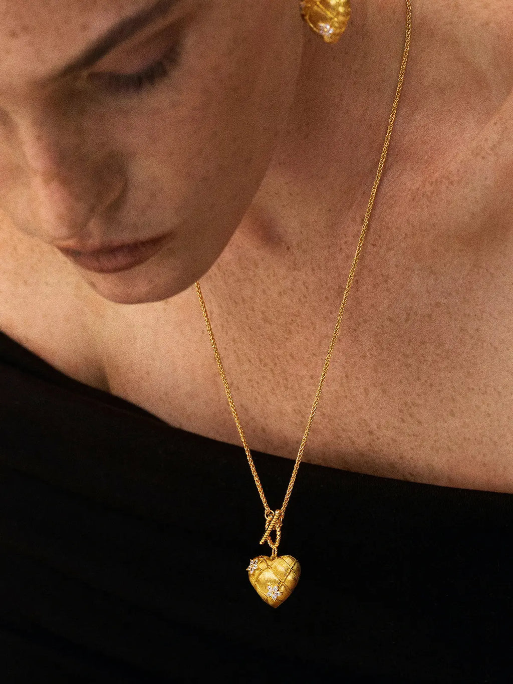 Close-up portrait of a model wearing the Tactilery Love Blossom Necklace with gaze downward, the large matte brushed gold quilted diamond-grid puffy heart pendant with marquise zircon blossom flower accents and twisted gold rope OT toggle clasp hanging at the décolletage on a fine polished gold wheat chain, against a dark background wearing a black top with short light hair and matching gold blossom drop earrings visible at the ear