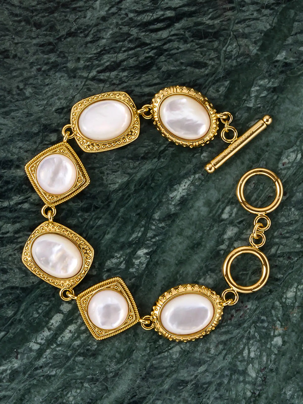 Tactilery Moonlight Box Mother of Pearl Bracelet in polished gold displayed lying flat on a dark green marble surface, the full link bracelet shown face-on with seven alternating cushion-square and oval polished gold granulated milgrain border frames each set with a large smooth white mother-of-pearl cabochon, connected by small gold jump rings, with the polished gold toggle bar-and-ring clasp visible at right