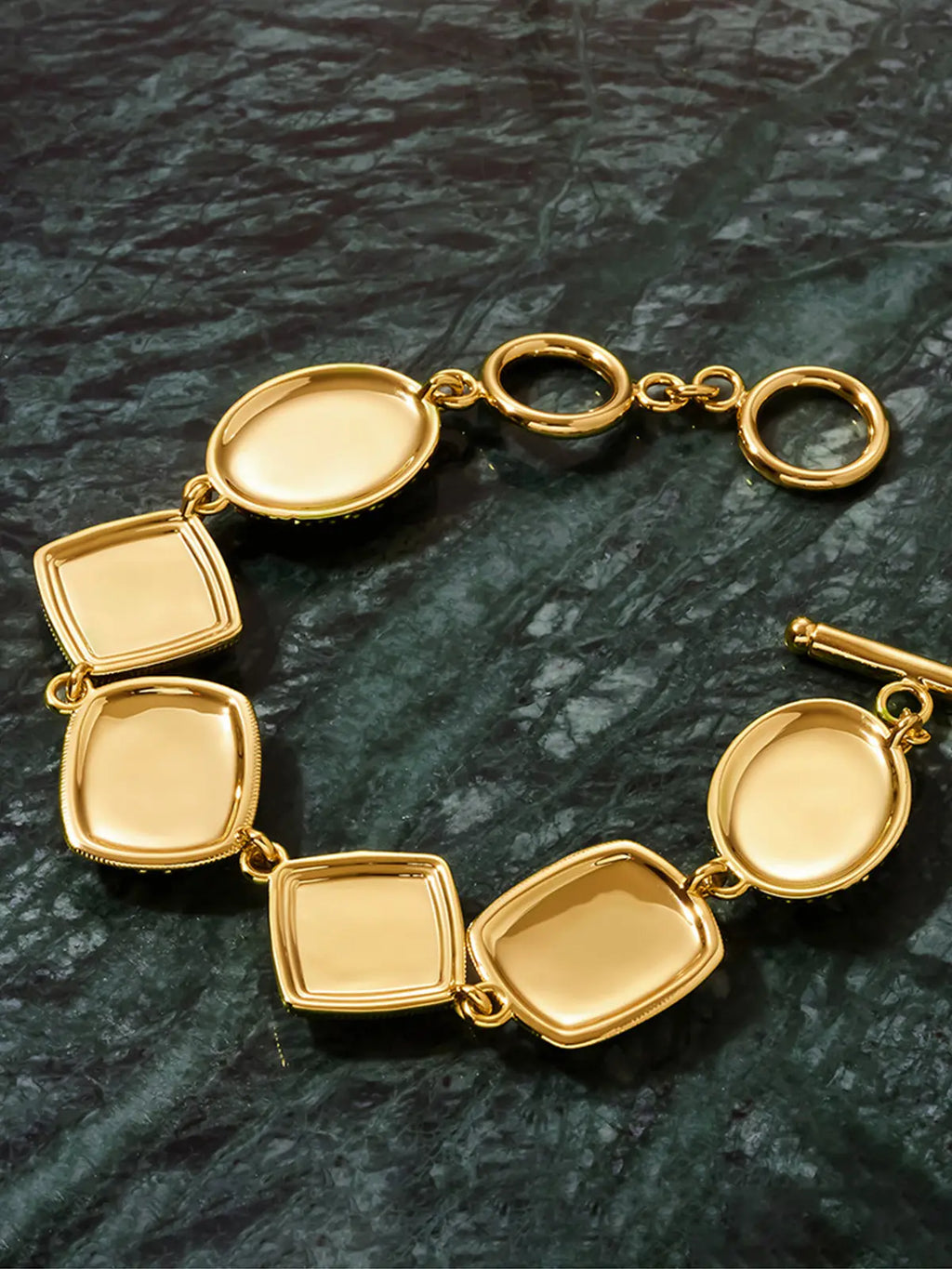 Tactilery Moonlight Box Mother of Pearl Bracelet in polished gold displayed lying flat on a dark green marble surface showing the back of the bracelet, the seven alternating cushion-square and oval polished gold frames revealed as smooth shallow open-back bezels without stone settings, showing the clean polished gold interior of each frame and the toggle bar-and-ring clasp at right