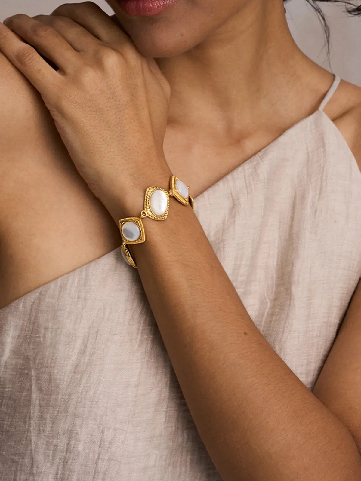 Close-up of a model's wrist wearing the Tactilery Moonlight Box Mother of Pearl Bracelet, the alternating cushion-square and oval polished gold milgrain border links each set with a large smooth white mother-of-pearl cabochon sitting at the wrist, against a warm taupe background wearing a light linen one-shoulder top with dark hair pulled back