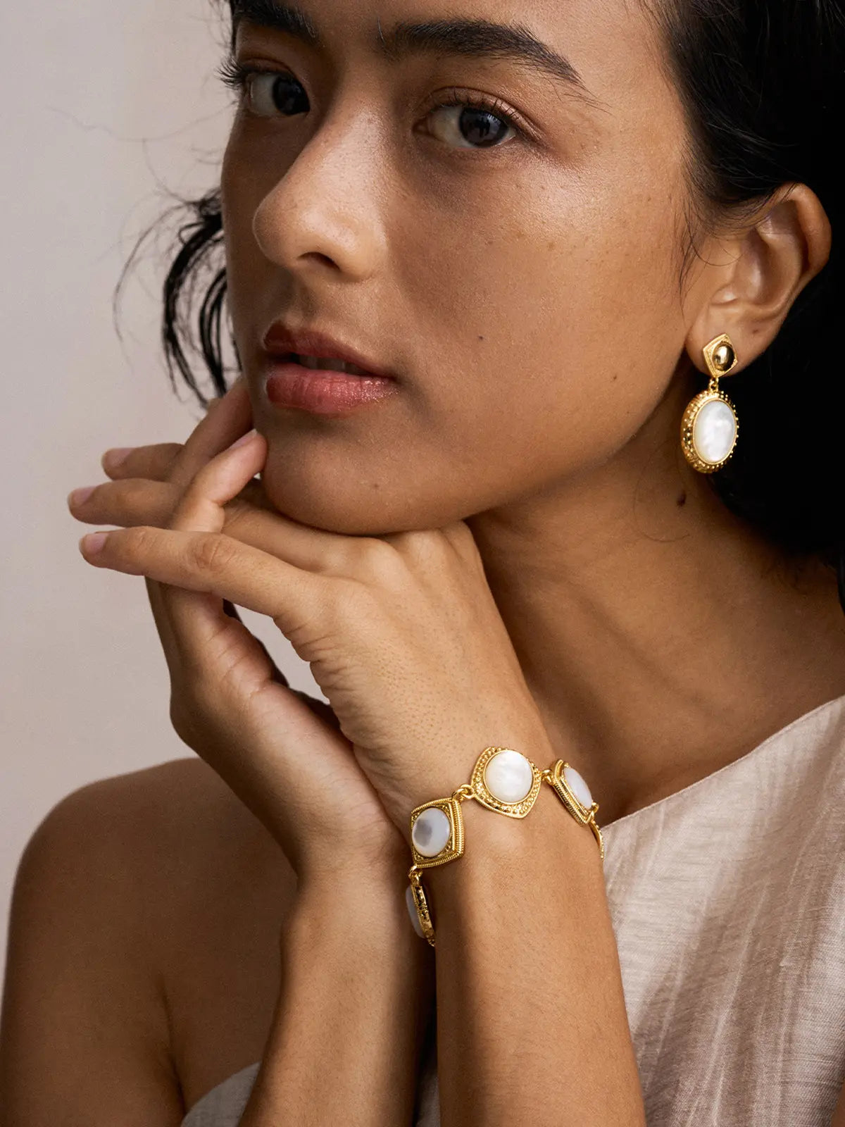 Three-quarter portrait of a model wearing the Tactilery Moonlight Box Mother of Pearl Bracelet on the wrist with hands raised to chin and direct gaze, the alternating cushion-square and oval polished gold milgrain border links each set with a large smooth white mother-of-pearl cabochon visible at the wrist, paired with matching moonlight box mother-of-pearl drop earrings, against a warm taupe background wearing a light linen one-shoulder top with dark hair pulled back