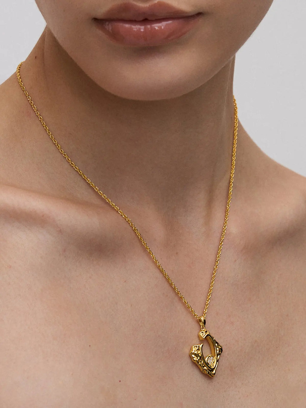 Close-up of a model's décolletage wearing the Tactilery Baroque Frame Necklace, the abstract diamond-shaped open-frame polished gold pendant with irregular molten border and small pavé zircon flower cluster accent hanging at the neckline on a polished gold twisted rope chain, against a cool grey background wearing a black strapless top
