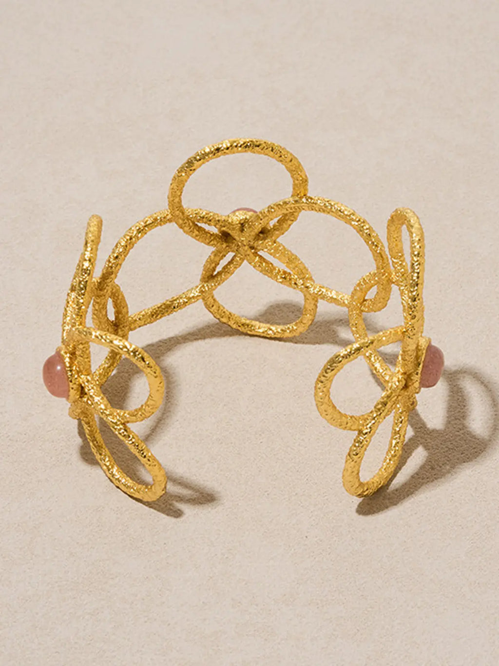 Tactilery Openwork Flower Cuff Bracelet in matte sand-textured gold displayed upright on a warm cream surface, the open C-cuff shown from the back revealing the full width of the interlocking looped gold wire lattice with three overlapping five-petal flower outlines and three pink rose quartz cabochon accents, the open cuff gap visible at center