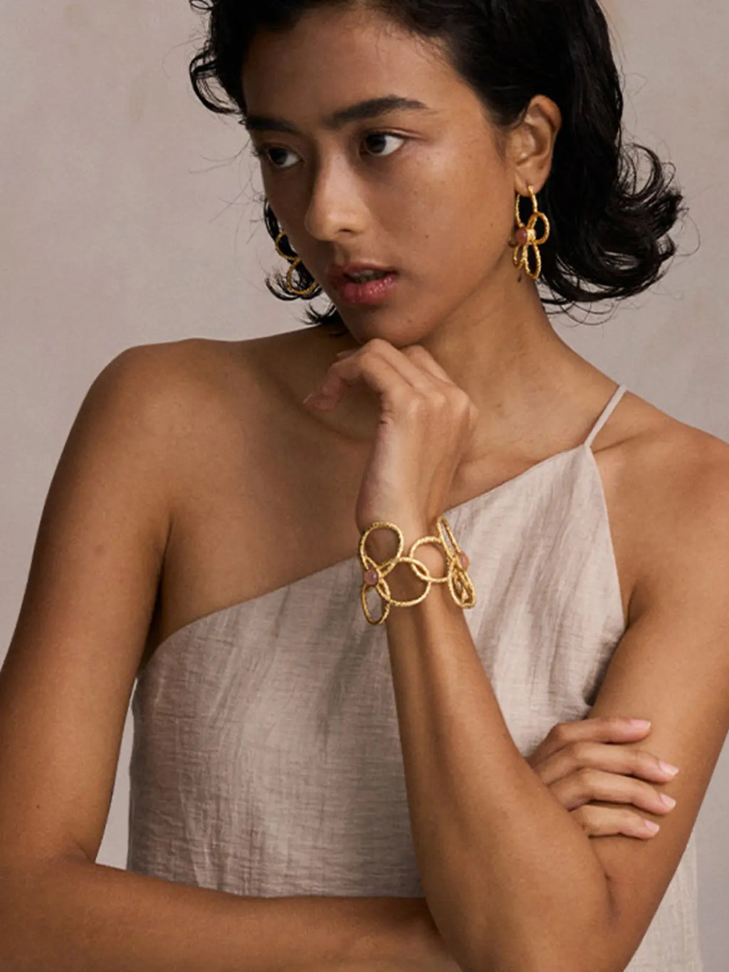 Three-quarter portrait of a model wearing the Tactilery Openwork Flower Cuff Bracelet on the wrist with hand raised to chin, the wide matte sand-textured gold wire lattice cuff with three overlapping five-petal flower outlines and pink rose quartz cabochon accents sitting at the wrist, paired with matching gold openwork loop drop earrings, against a warm taupe background wearing a light linen one-shoulder top with short dark wavy hair