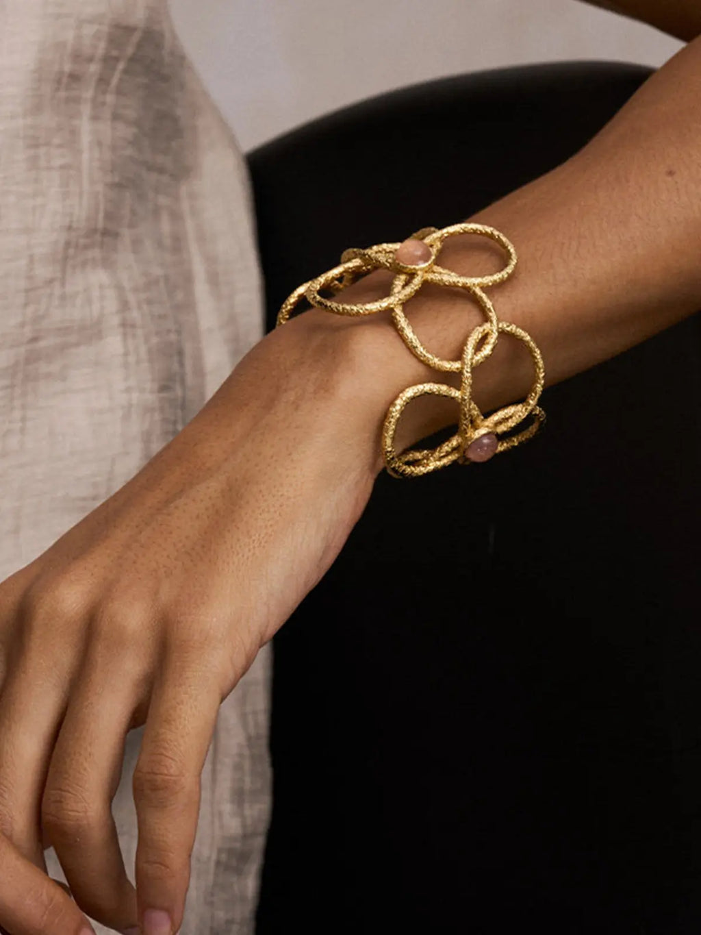 Close-up of a model's wrist wearing the Tactilery Openwork Flower Cuff Bracelet, the wide matte sand-textured gold wire lattice cuff with three overlapping five-petal flower outlines and pink rose quartz cabochon accents filling the frame, against a dark background wearing a light linen top
