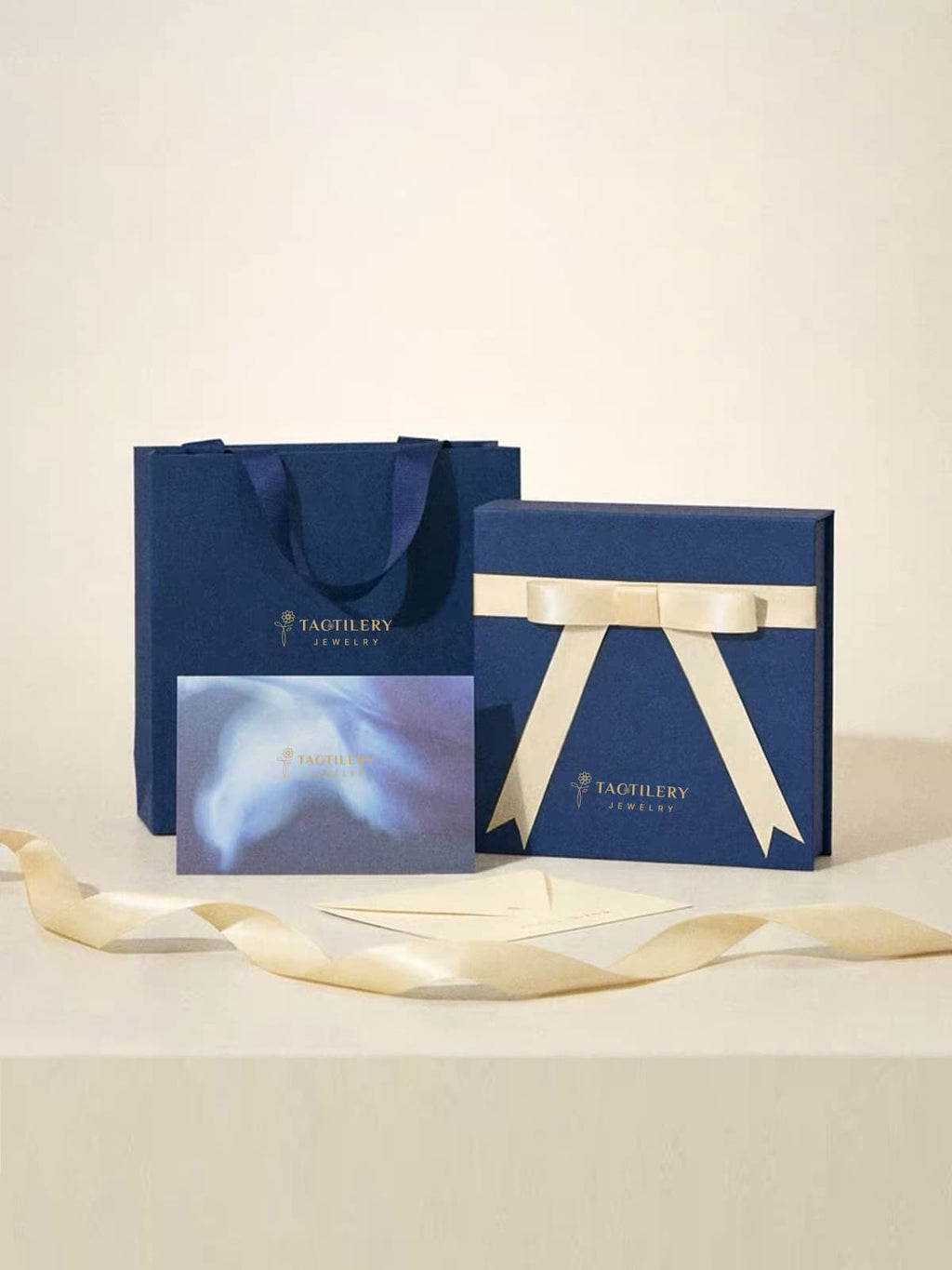 A Tagilery jewelry gift set featuring a navy blue shopping bag, a navy blue presentation box tied with a cream ribbon, a matching card, and an envelope on a light beige background.