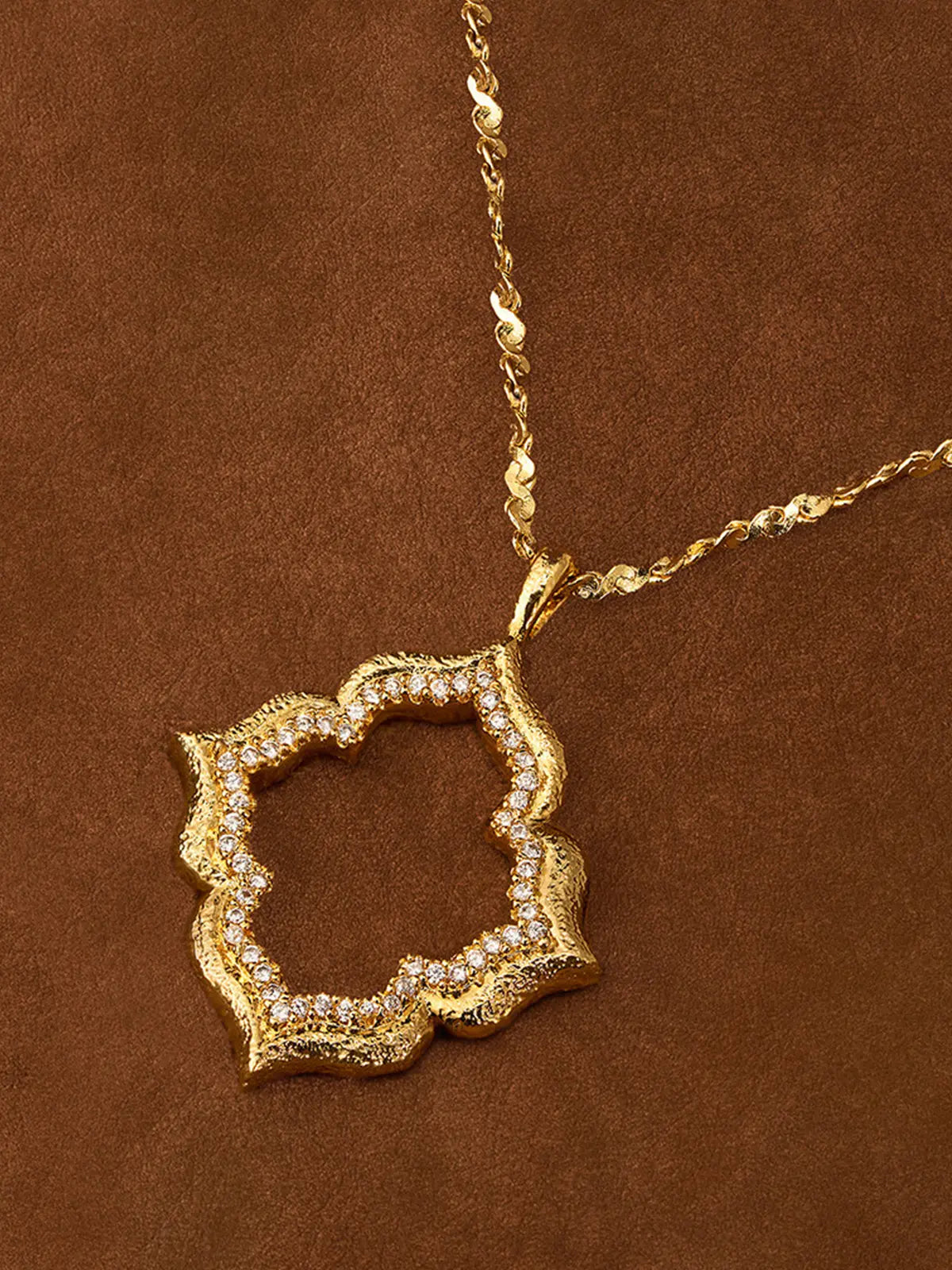 Extreme close-up of the Tactilery Persimmon Calyx Openwork Necklace pendant on a white surface, the large open-frame four-lobe persimmon calyx flower with matte sand-textured gold border and deeply scalloped continuous pavé zircon inner edge in sharp detail, with the fine polished gold twisted petal-link chain bail visible above