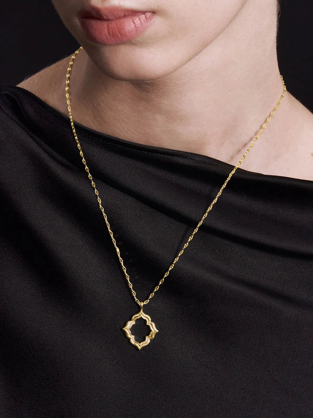 Three-quarter portrait of a model wearing the Tactilery Persimmon Calyx Openwork Necklace with gaze downward, the large open-frame four-lobe persimmon calyx flower pendant with matte sand-textured gold border and scalloped pavé zircon inner edge visible at the décolletage on a fine polished gold twisted petal-link chain, against a warm taupe background wearing a white draped top with dark hair in a low bun