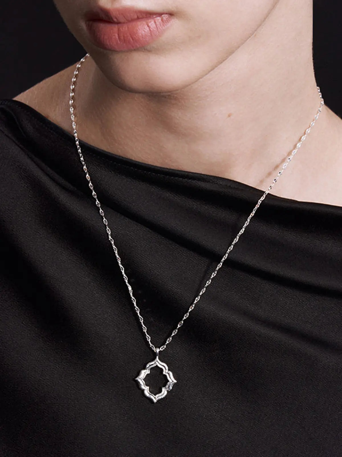 Three-quarter portrait of a model wearing the Tactilery Persimmon Calyx Openwork Necklace with gaze to the side, the large open-frame four-lobe persimmon calyx flower pendant with matte sand-textured gold border and scalloped pavé zircon inner edge visible at the décolletage on a fine polished gold twisted petal-link chain, against a warm taupe background wearing a white draped top with dark hair pulled back