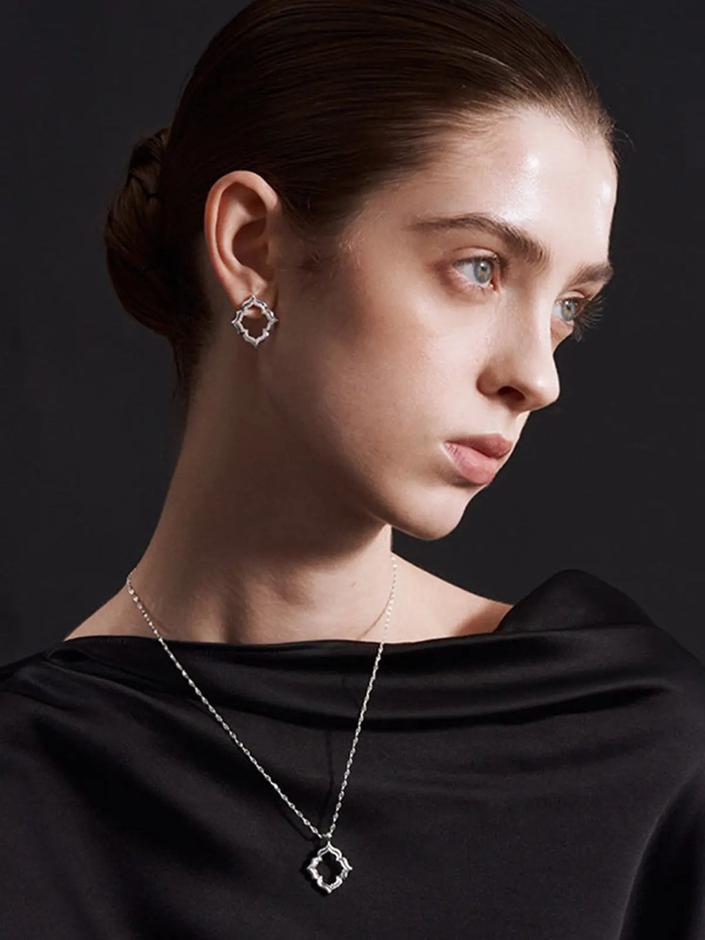 Close-up of a model's décolletage wearing the Tactilery Persimmon Calyx Openwork Necklace from a slightly elevated angle, the large open-frame four-lobe persimmon calyx flower pendant with matte sand-textured gold border and scalloped pavé zircon inner edge sitting at the neckline on a fine polished gold twisted petal-link chain, against a warm taupe background wearing a white draped top