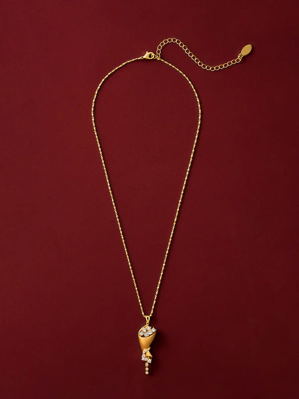 Tactilery Bouquet Necklace in matte brushed gold displayed laid out flat on a deep red surface showing the full necklace length, the polished gold twisted rope chain extending the full length with the lobster clasp and oval tag charm extension chain at top and the sculptural cone bouquet pendant at bottom