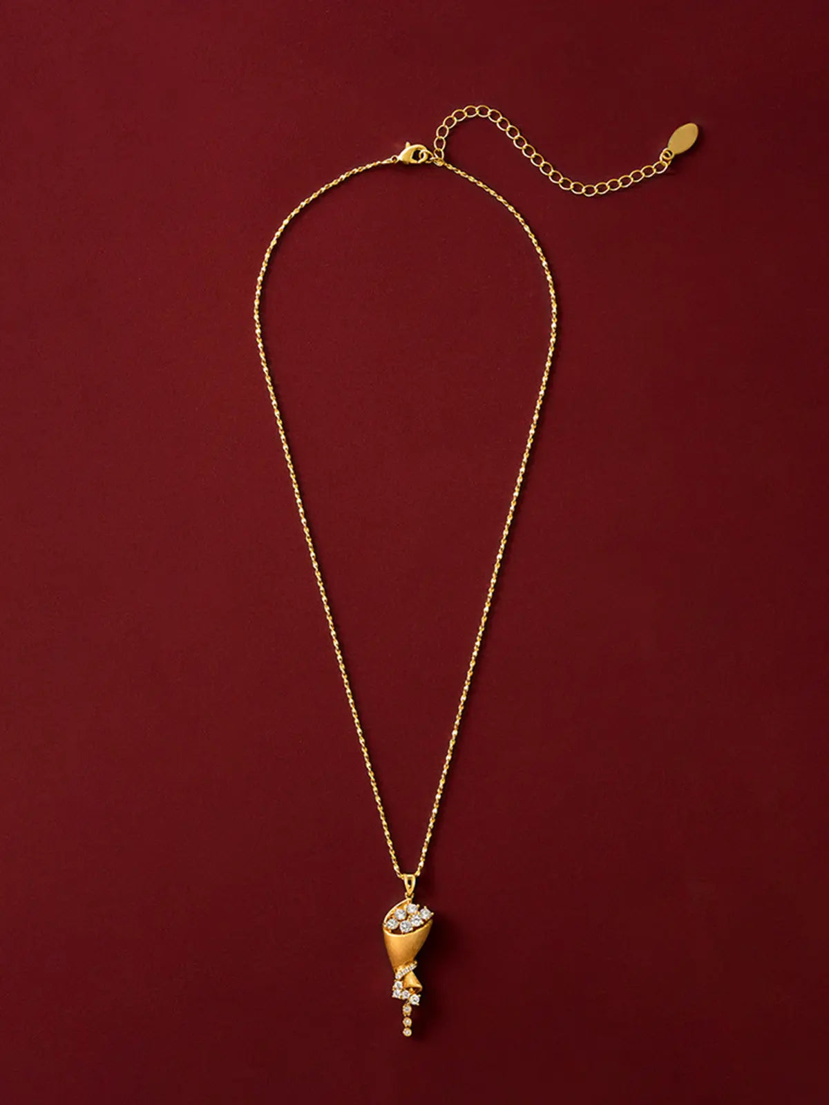 Tactilery Bouquet Necklace in matte brushed gold displayed laid out flat on a deep red surface showing the full necklace length, the polished gold twisted rope chain extending the full length with the lobster clasp and oval tag charm extension chain at top and the sculptural cone bouquet pendant at bottom