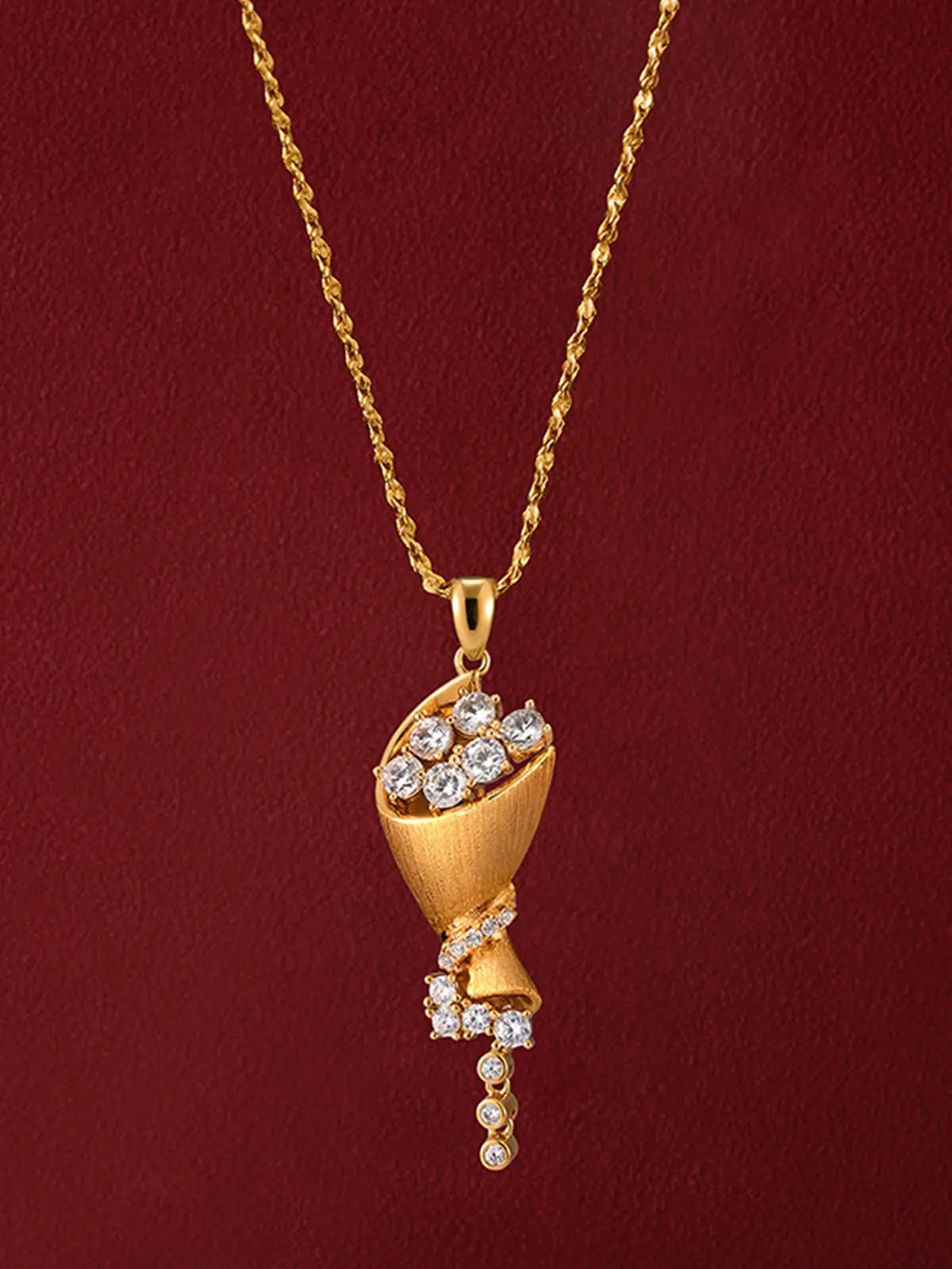 Tactilery Bouquet Necklace in matte brushed gold displayed on a deep red surface, the sculptural cone bouquet pendant with cluster of seven prong-set large round zircons at top, pavé zircon twisted ribbon wrap spiraling around the lower cone body, and trailing spray of three prong-set graduating round zircons and three bezel-set round zircon drops on fine gold wire stems, suspended from a polished gold twisted rope chain