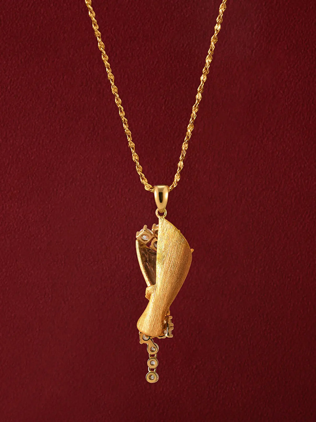 Tactilery Bouquet Necklace in matte brushed gold displayed on a deep red surface showing the back of the pendant, the matte brushed gold cone-shaped wrap with fine horizontal brushed texture shown from the back — the smooth brushed gold back face of the cone and the pavé zircon twisted ribbon wrap visible from behind — with the polished gold twisted rope chain and bail above