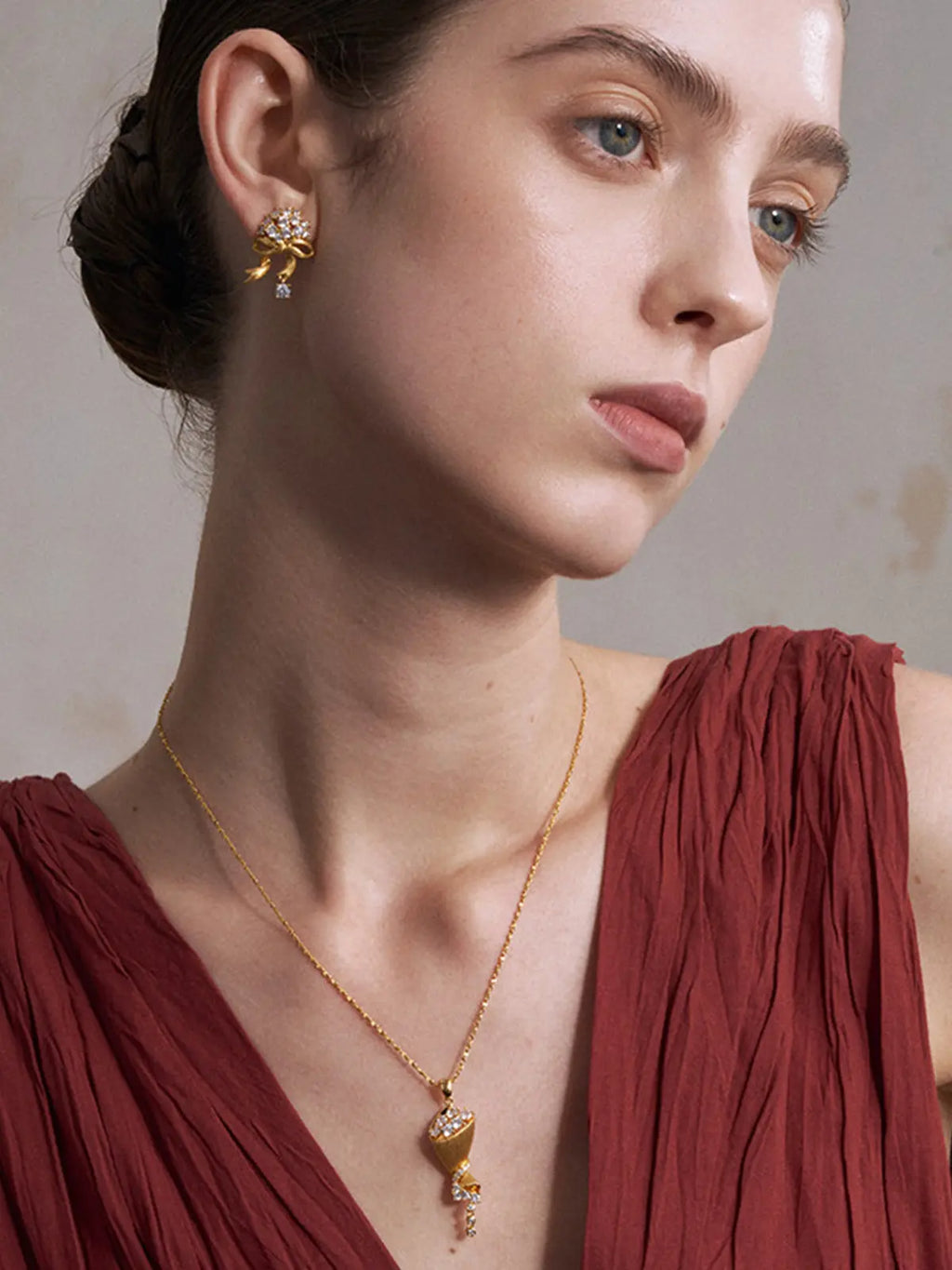 A close-up of a woman wearing a Tactilery bouquet necklace with a sculptural pendant and matching gold earrings, paired with a draped burgundy top against a neutral studio background.