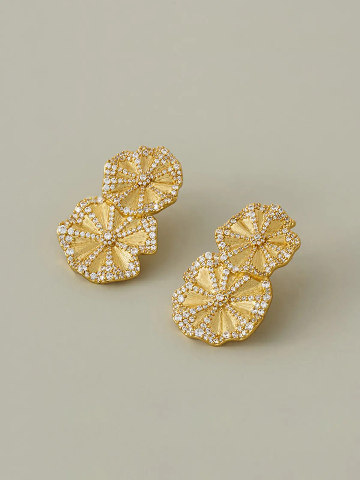 Pair of Tactilery Rain Lotus Leaf Stud Earrings in matte brushed gold displayed at an angle on a warm sage green surface, one showing the full front face of the double lotus leaf drop with smaller scalloped pavé zircon border lotus leaf at top and larger scalloped pavé zircon border lotus leaf below with radiating ribbed vein texture, one showing the three-dimensional depth of the stacked leaf form