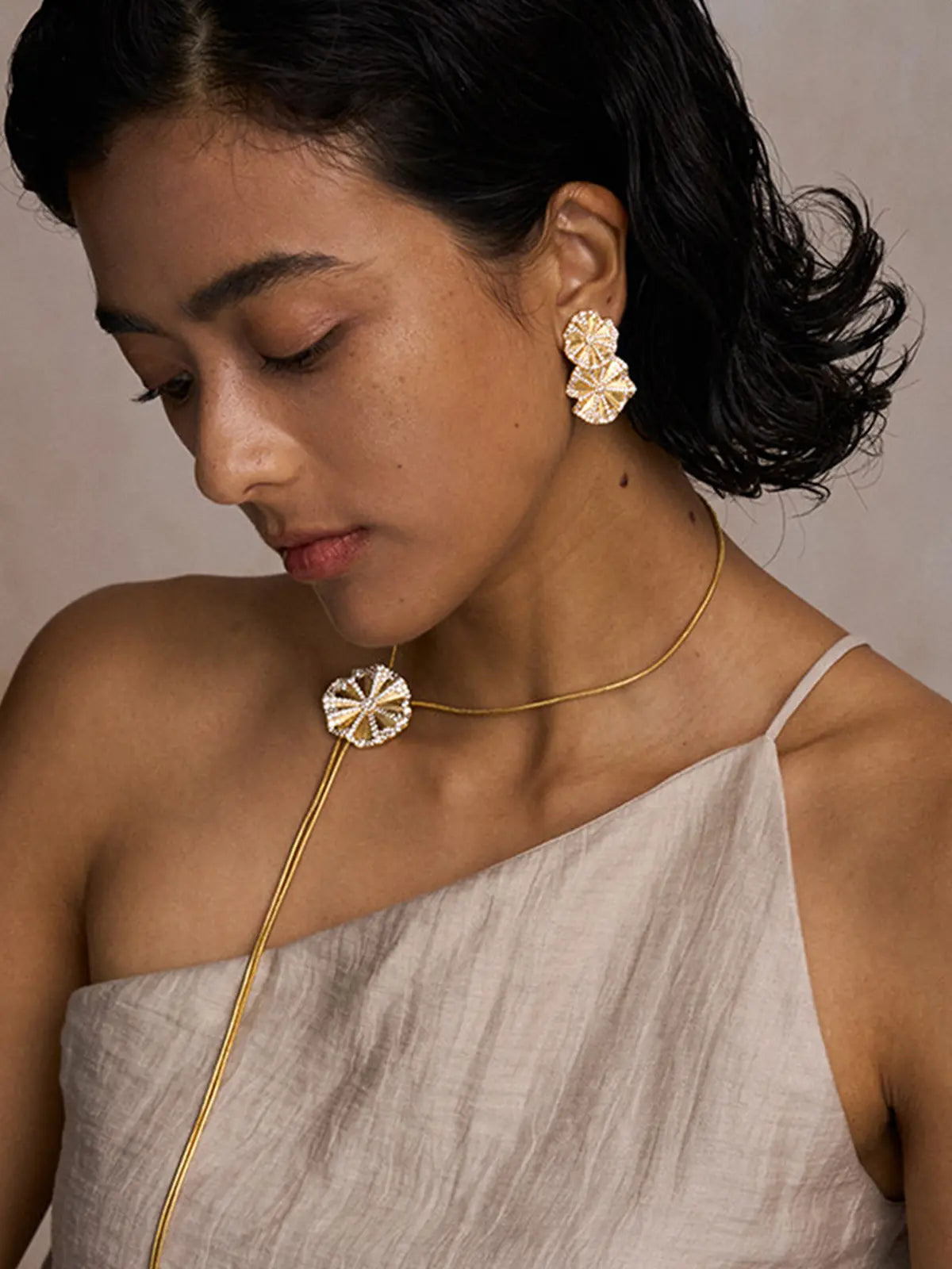 Three-quarter portrait of a model wearing the Tactilery Rain Lotus Leaf Stud Earrings with gaze downward and hand raised to hair, the double lotus leaf drop with smaller and larger scalloped pavé zircon border lotus leaves dangling at the lobe, paired with a matching Rain Lotus Leaf pendant on a long gold snake chain necklace, against a warm grey background wearing a light linen one-shoulder top with short dark wavy hair