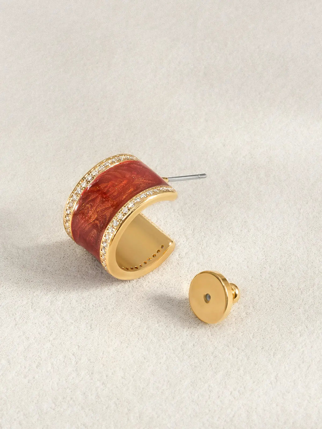 Single Tactilery Red Glaze Zircon Hoop Earring in polished gold displayed at an angle on a warm grey surface with the gold disc butterfly back removed and placed beside it, showing the silver post pin at top and the three-dimensional depth of the wide chunky C-hoop with deep vermilion red crinkled enamel glaze panel bordered above and below by continuous pavé zircon rows, revealing the smooth polished gold inner band
