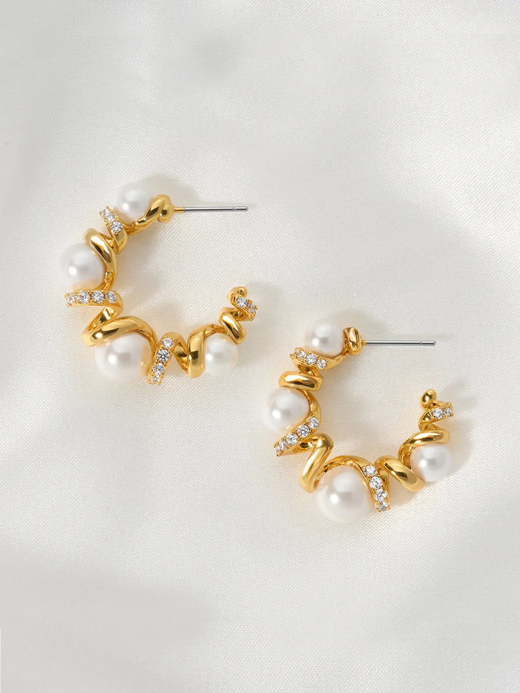Pair of Tactilery Spiral Twist Pearl Hoop Earrings in polished gold displayed lying flat on a white fabric surface, both showing the face-on view of the C-hoop with five graduated white pearl spheres interspersed along the polished gold spiral twist wire with pavé zircon bar accents at each junction, with silver post pins visible at top