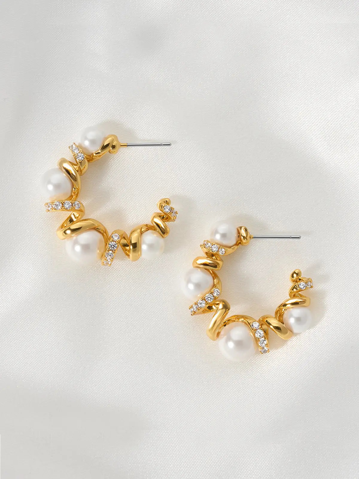 Pair of Tactilery Spiral Twist Pearl Hoop Earrings in polished gold displayed lying flat on a white fabric surface, both showing the face-on view of the C-hoop with five graduated white pearl spheres interspersed along the polished gold spiral twist wire with pavé zircon bar accents at each junction, with silver post pins visible at top