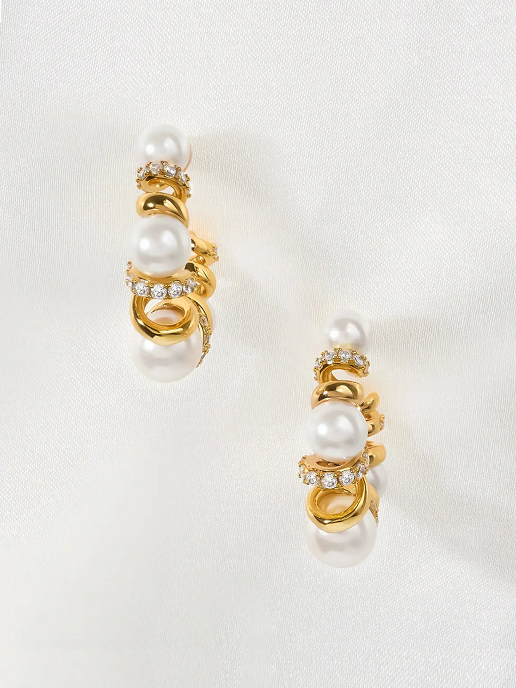 Pair of Tactilery Spiral Twist Pearl Hoop Earrings in polished gold displayed upright on a white fabric surface, both in profile showing the three-dimensional depth of the spiral twist wire construction with five graduated white pearl spheres and pavé zircon bar accents, with silver post pins visible at top