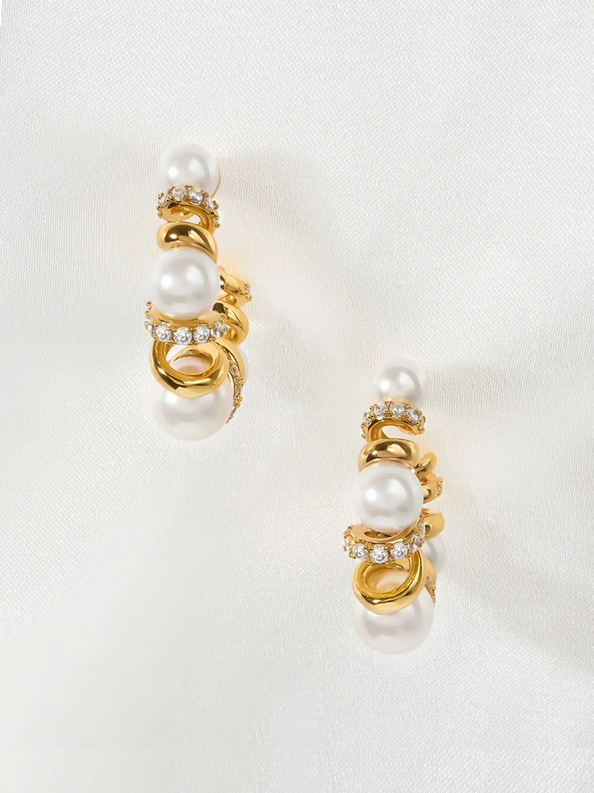 Pair of Tactilery Spiral Twist Pearl Hoop Earrings in polished gold displayed upright on a white fabric surface, both in profile showing the three-dimensional depth of the spiral twist wire construction with five graduated white pearl spheres and pavé zircon bar accents, with silver post pins visible at top