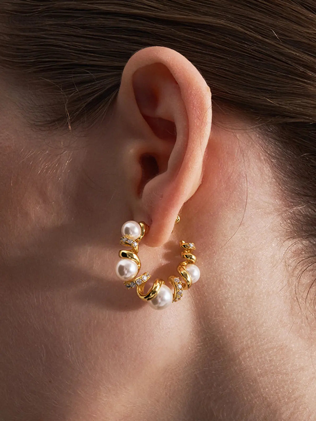 Extreme close-up profile of a model's ear wearing the Tactilery Spiral Twist Pearl Hoop Earrings, the large polished gold spiral twist C-hoop with five graduated white pearl spheres and pavé zircon bar accents sitting at the lobe, against a warm brown background with dark hair slicked back