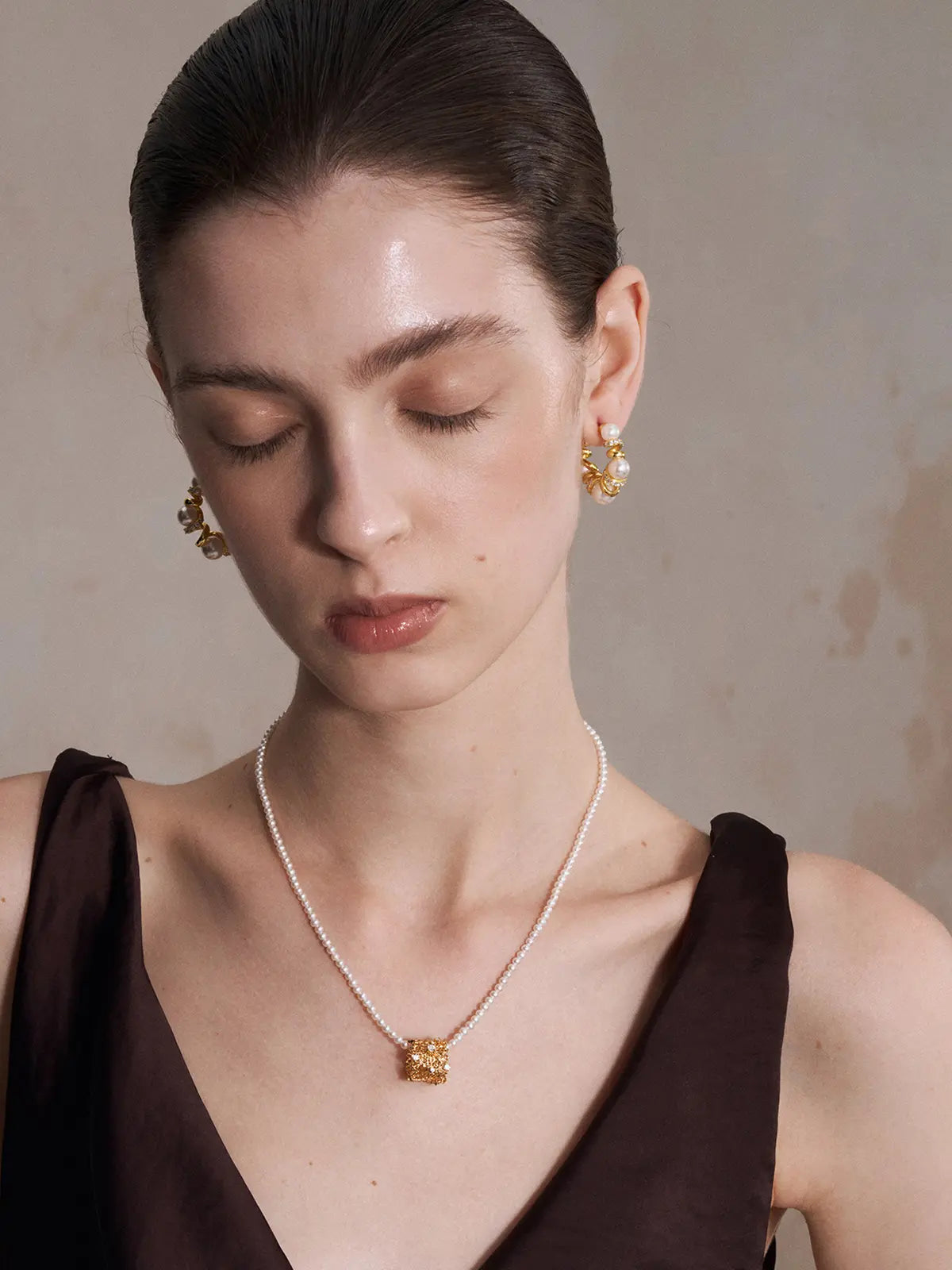 Three-quarter portrait of a model wearing the Tactilery Spiral Twist Pearl Hoop Earrings on both ears with gaze downward, the large polished gold spiral twist C-hoop with five graduated white pearl spheres and pavé zircon bar accents visible at both lobes, paired with a fine pearl strand necklace with a gold baroque pendant, against a warm taupe background wearing a dark brown sleeveless top with dark hair slicked back