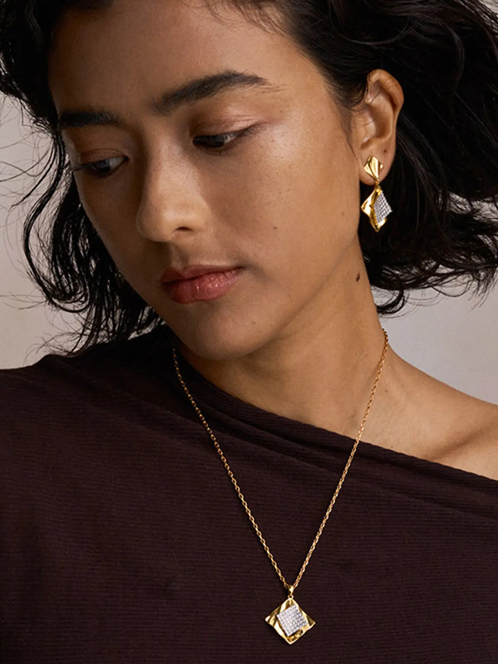 Three-quarter portrait of a model wearing the Tactilery Streamer Wave Diamond Necklace with gaze downward, the square diamond-shaped pendant with wave-rippled polished gold outer frame and central square pavé zircon panel hanging at the décolletage on a fine polished gold cable chain, paired with matching streamer wave diamond drop earrings with the same wave-rippled gold frame and pavé zircon panel, against a warm taupe background wearing a dark top with short dark wavy hair