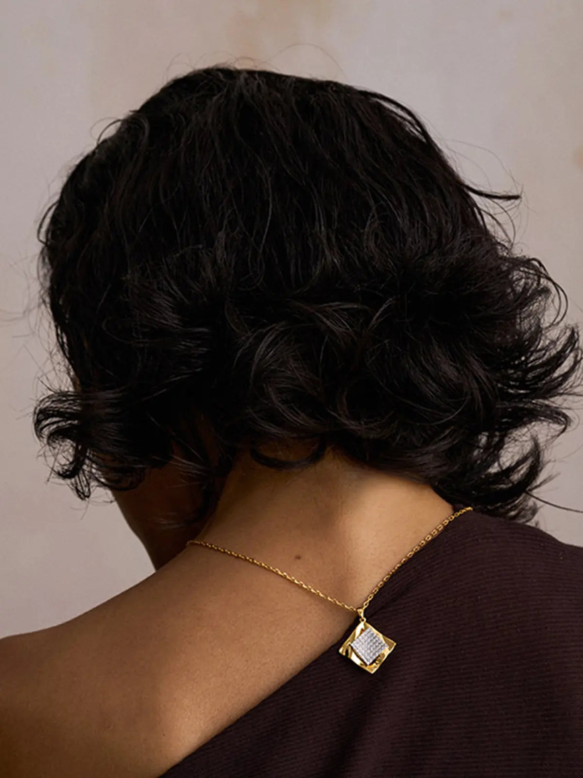Back-of-head portrait of a model wearing the Tactilery Streamer Wave Diamond Necklace, the square diamond-shaped pendant with wave-rippled polished gold outer frame and central square pavé zircon panel visible at the upper back neckline on a fine polished gold cable chain, against a warm taupe background wearing a dark one-shoulder top with short dark wavy hair