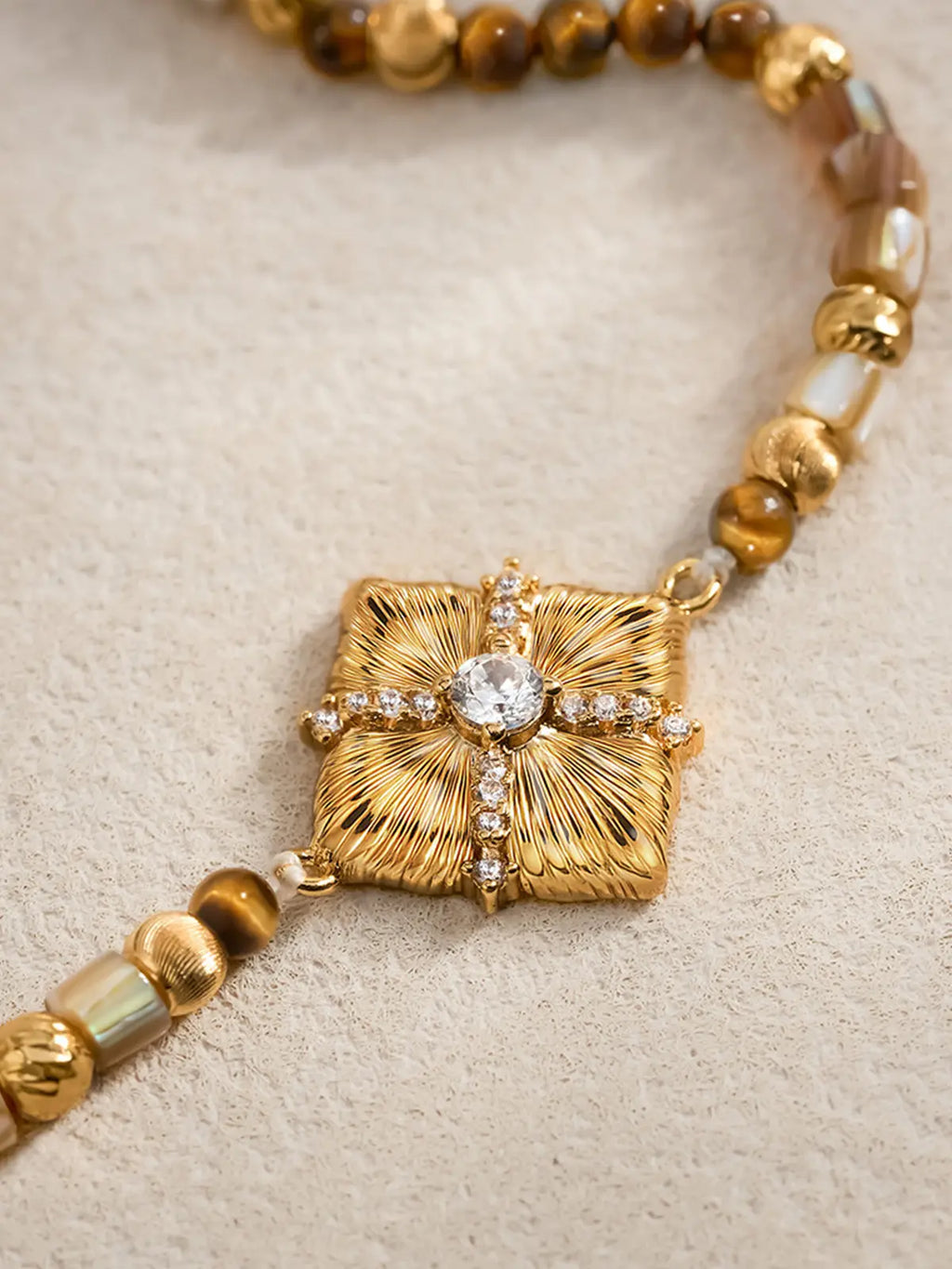 Extreme close-up of the Tactilery Sugar Cube Beaded Bracelet center pendant on a warm sand textured surface, the large square diamond-shaped polished gold pendant with deeply radiating sunburst line-engraved texture filling the frame — the central prong-set large round zircon and pavé zircon cross accent in sharp detail — flanked by caramel brown tiger eye round beads, translucent grey-brown rectangular tube beads, and polished gold ball spacer beads