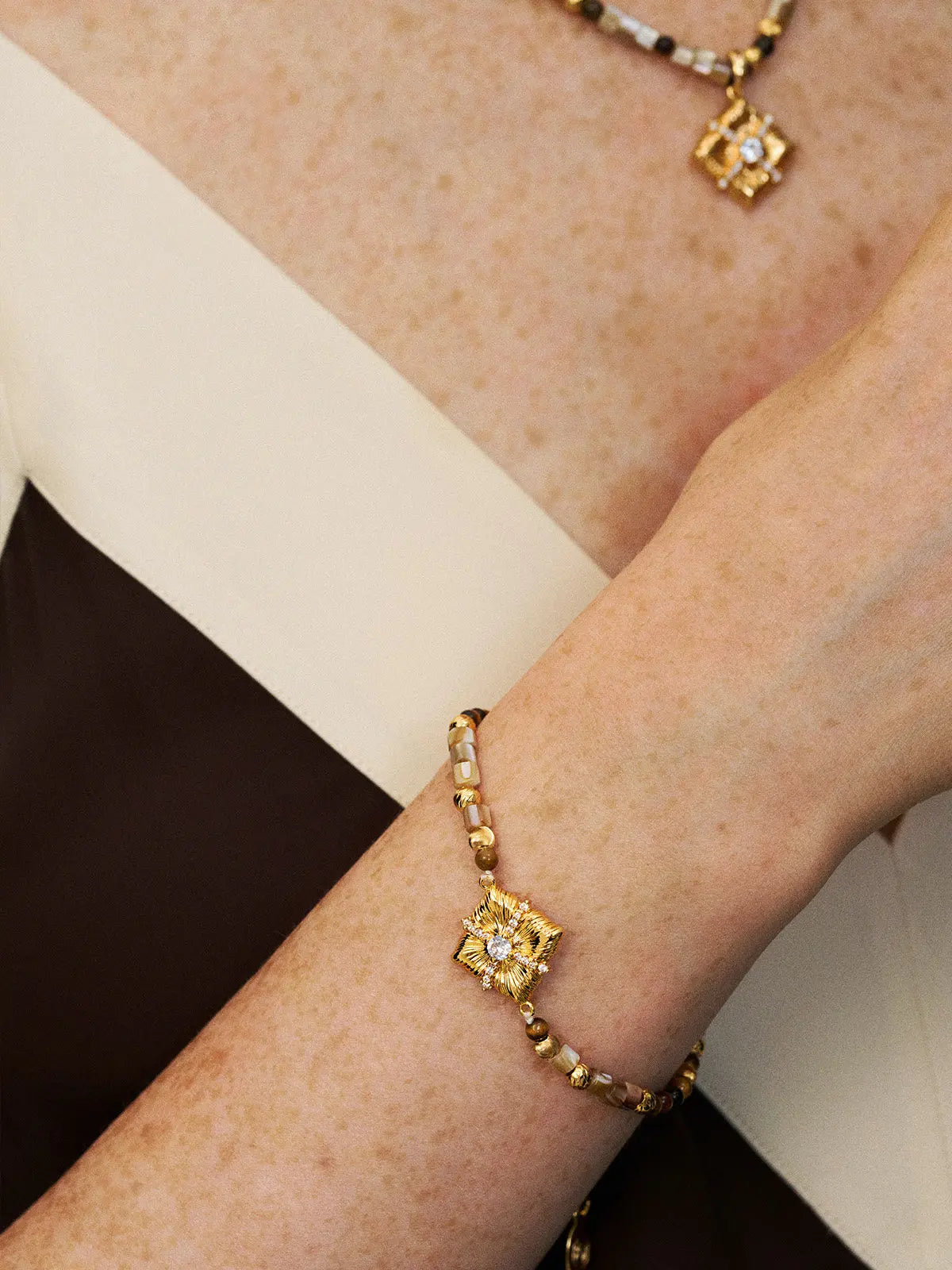Close-up of a model's wrist wearing the Tactilery Sugar Cube Beaded Bracelet, the beaded strand of alternating caramel brown tiger eye round beads, translucent grey-brown rectangular tube beads, and polished gold ball spacer beads with the large square diamond-shaped sunburst-engraved gold pendant and pavé zircon cross accent sitting at the wrist, with a matching sugar cube beaded necklace visible at the collarbone, against a warm cream and dark background wearing a dark top