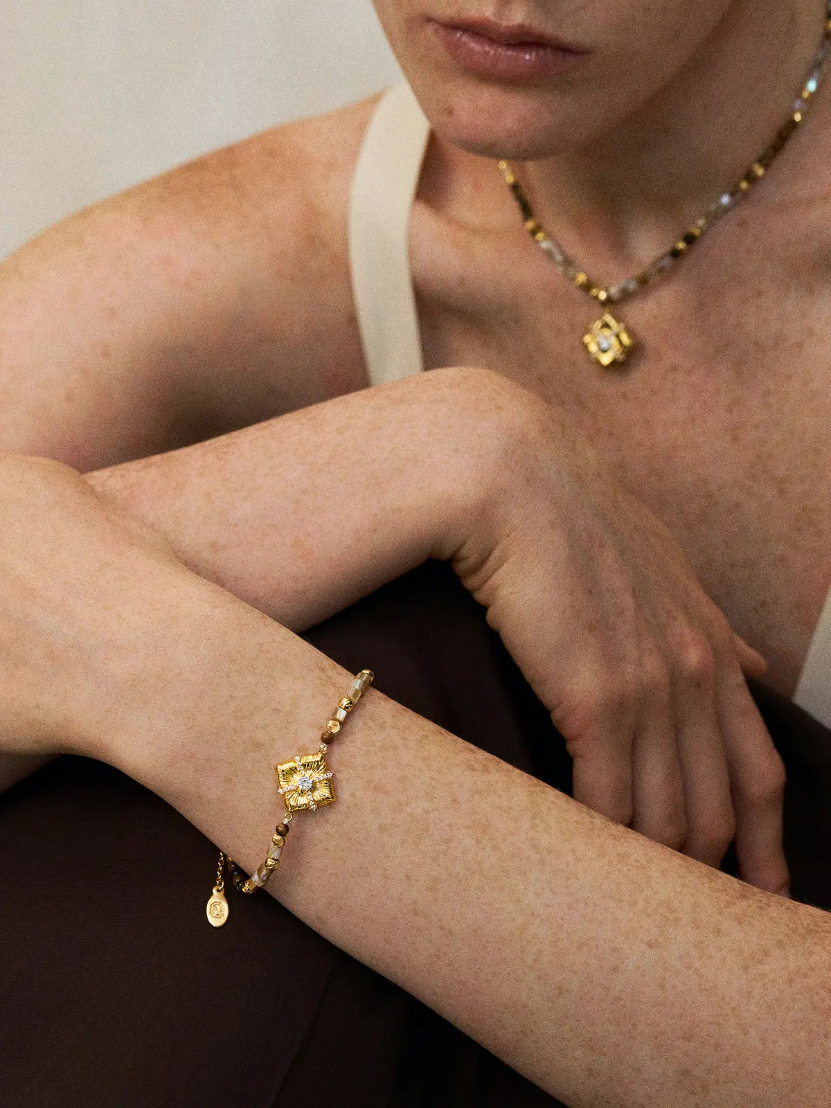 Three-quarter portrait of a model wearing the Tactilery Sugar Cube Beaded Bracelet on the wrist with arms crossed and gaze downward, the beaded bracelet with the large square diamond-shaped sunburst-engraved gold pendant and pavé zircon cross accent visible at the wrist, with a matching sugar cube beaded necklace visible at the collarbone, against a warm taupe background wearing a white sleeveless top