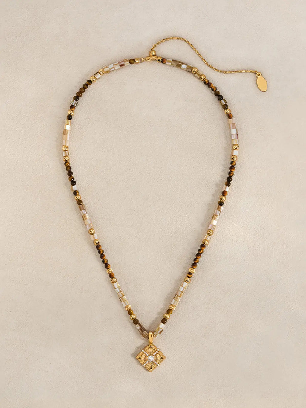 Tactilery Sugar Cube Beaded Necklace in polished gold displayed laid out flat on a warm cream surface showing the full necklace length, the complete beaded strand of alternating mother-of-pearl tile, tiger's eye, and polished gold textured beads with the lobster clasp and polished gold oval tag charm extension chain at top and the square diamond-shaped sunburst pendant at bottom center