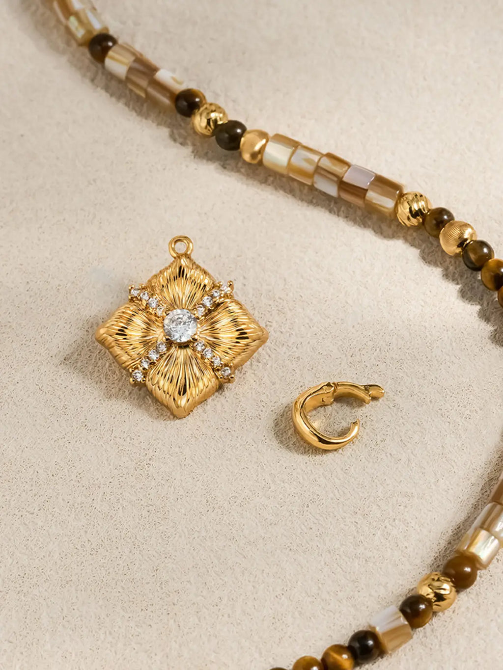 Flat-lay detail of the Tactilery Sugar Cube Beaded Necklace on a warm cream surface, the square diamond-shaped polished gold sunburst pendant with four deeply radiating line-engraved petal-like lobes, central prong-set large round zircon, and pavé zircon cross accent shown alongside the beaded strand and a small polished gold C-hoop earring, the bead pattern of alternating mother-of-pearl tile, tiger's eye, and polished gold textured beads clearly visible