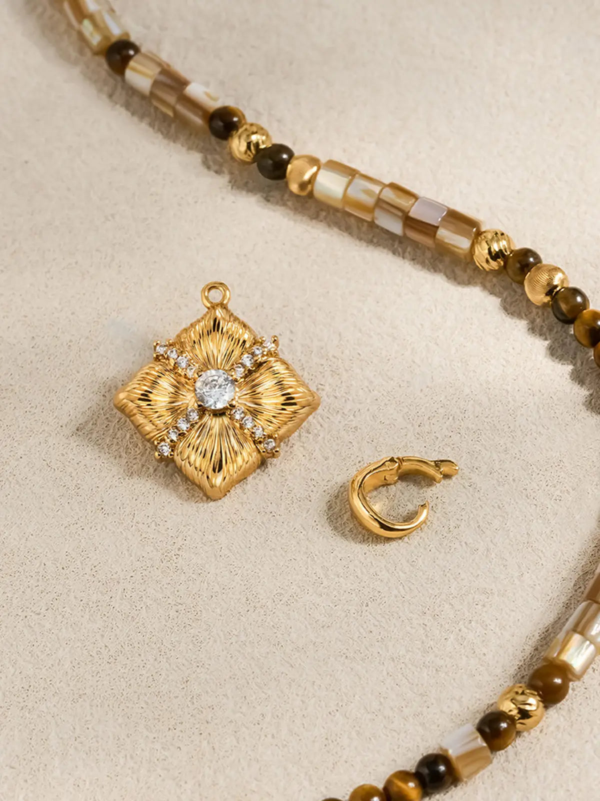 Flat-lay detail of the Tactilery Sugar Cube Beaded Necklace on a warm cream surface, the square diamond-shaped polished gold sunburst pendant with four deeply radiating line-engraved petal-like lobes, central prong-set large round zircon, and pavé zircon cross accent shown alongside the beaded strand and a small polished gold C-hoop earring, the bead pattern of alternating mother-of-pearl tile, tiger's eye, and polished gold textured beads clearly visible