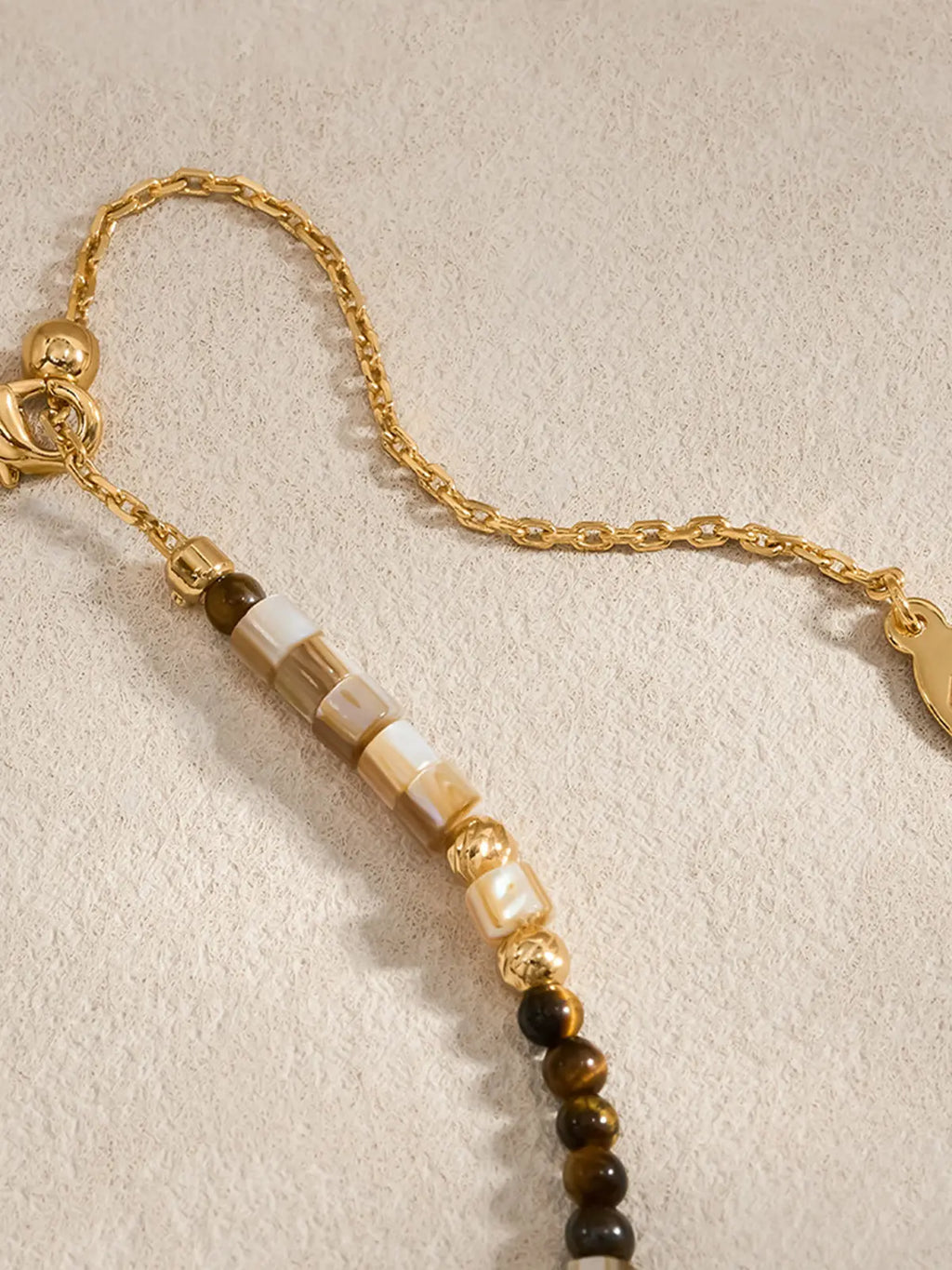 Extreme close-up of the Tactilery Sugar Cube Beaded Necklace clasp end on a warm cream surface, the polished gold lobster clasp and polished gold cable chain extension with oval tag charm visible at top, with the alternating mother-of-pearl tile, tiger's eye, and polished gold textured bead pattern of the strand shown in sharp detail below