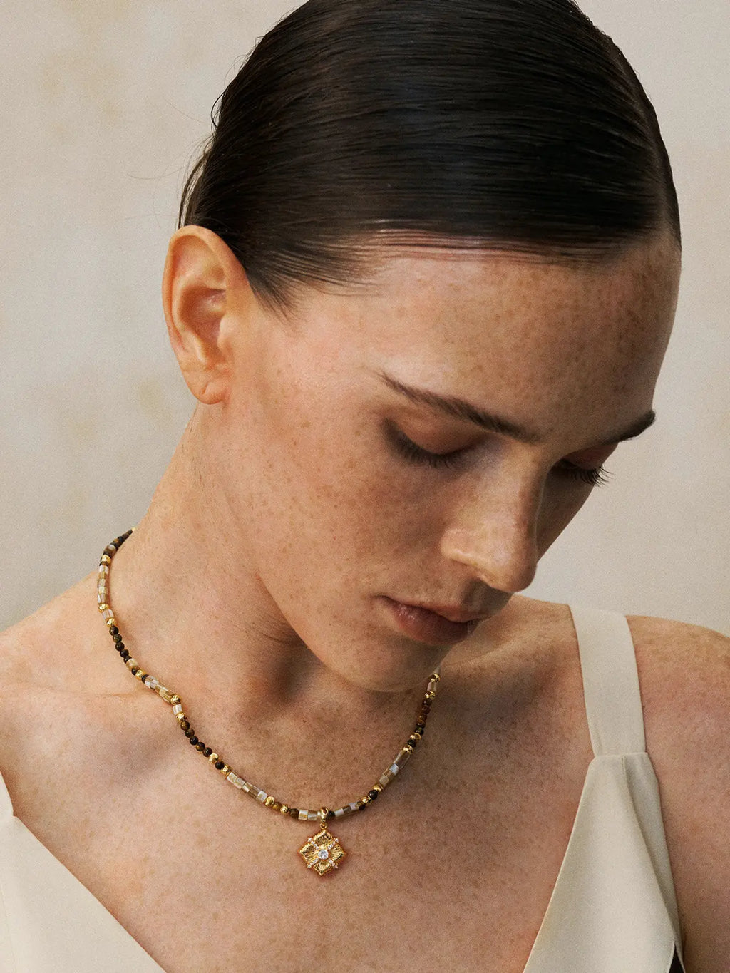 Three-quarter portrait of a model wearing the Tactilery Sugar Cube Beaded Necklace with gaze downward, the beaded strand of alternating mother-of-pearl tile, tiger's eye, and polished gold textured beads with the square diamond-shaped sunburst pendant and central prong-set zircon sitting at the collarbone, against a warm taupe background wearing a white and black V-neck top with dark hair slicked back