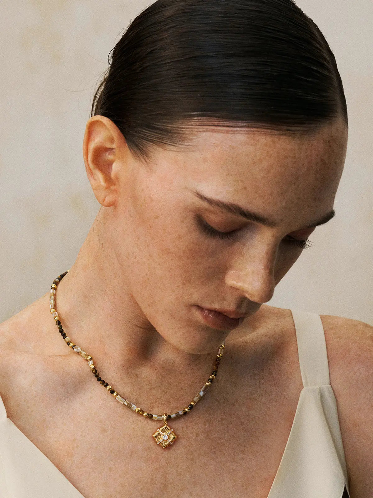 Three-quarter portrait of a model wearing the Tactilery Sugar Cube Beaded Necklace with gaze downward, the beaded strand of alternating mother-of-pearl tile, tiger's eye, and polished gold textured beads with the square diamond-shaped sunburst pendant and central prong-set zircon sitting at the collarbone, against a warm taupe background wearing a white and black V-neck top with dark hair slicked back