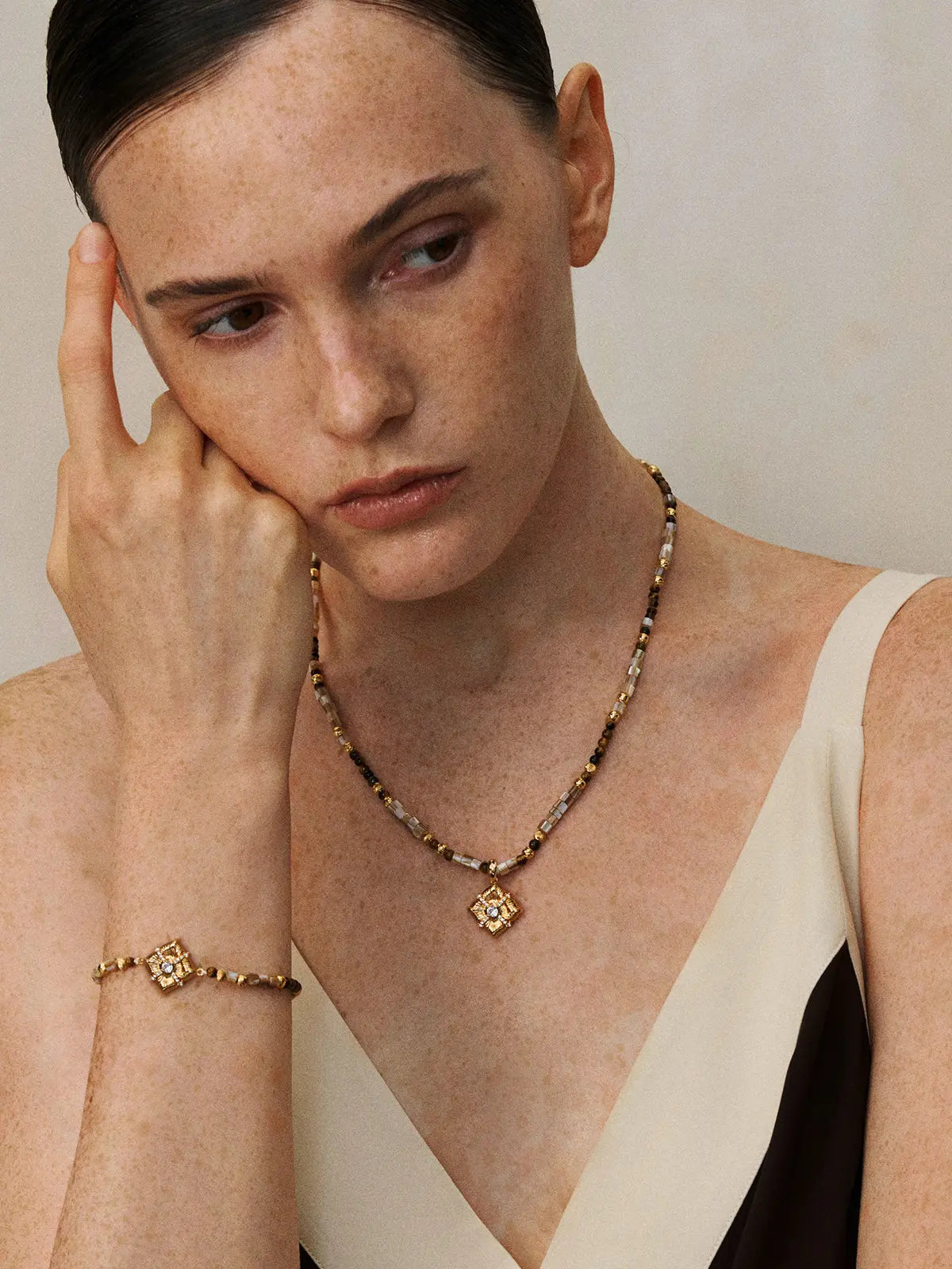 Three-quarter portrait of a model wearing the Tactilery Sugar Cube Beaded Necklace with hand raised to cheek and gaze downward, the beaded strand of alternating mother-of-pearl tile, tiger's eye, and polished gold textured beads with the square diamond-shaped sunburst pendant and central prong-set zircon sitting at the collarbone, paired with a matching sugar cube beaded bracelet on the wrist, against a warm taupe background wearing a white and black V-neck top with dark hair slicked back
