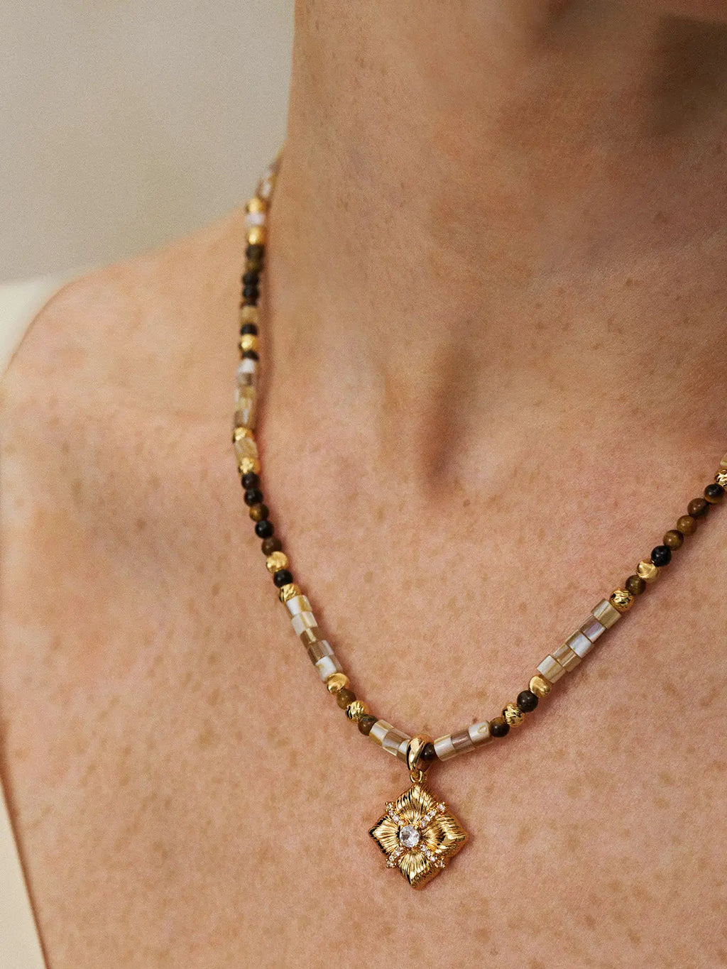 Close-up of a model's neck and décolletage wearing the Tactilery Sugar Cube Beaded Necklace, the beaded strand of alternating mother-of-pearl tile, tiger's eye, and polished gold textured beads with the square diamond-shaped sunburst pendant and central prong-set zircon clearly visible at the collarbone, against a warm taupe background wearing a white and black V-neck top
