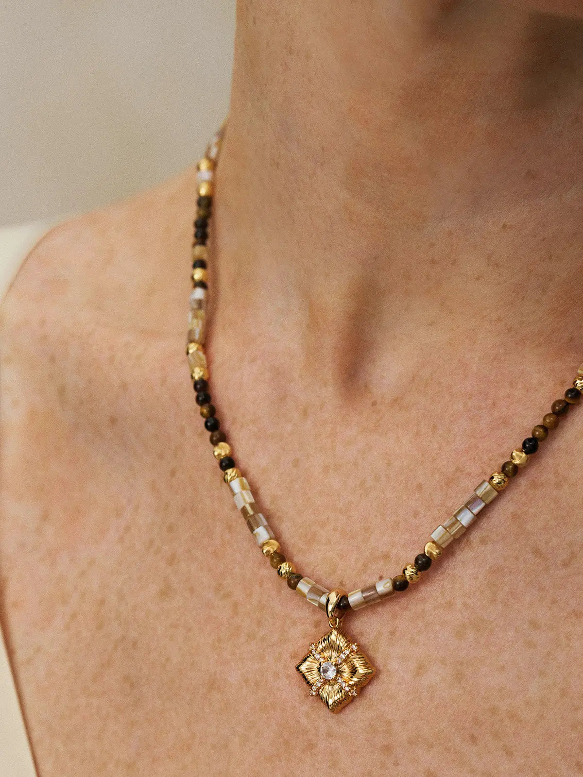Close-up of a model's neck and décolletage wearing the Tactilery Sugar Cube Beaded Necklace, the beaded strand of alternating mother-of-pearl tile, tiger's eye, and polished gold textured beads with the square diamond-shaped sunburst pendant and central prong-set zircon clearly visible at the collarbone, against a warm taupe background wearing a white and black V-neck top