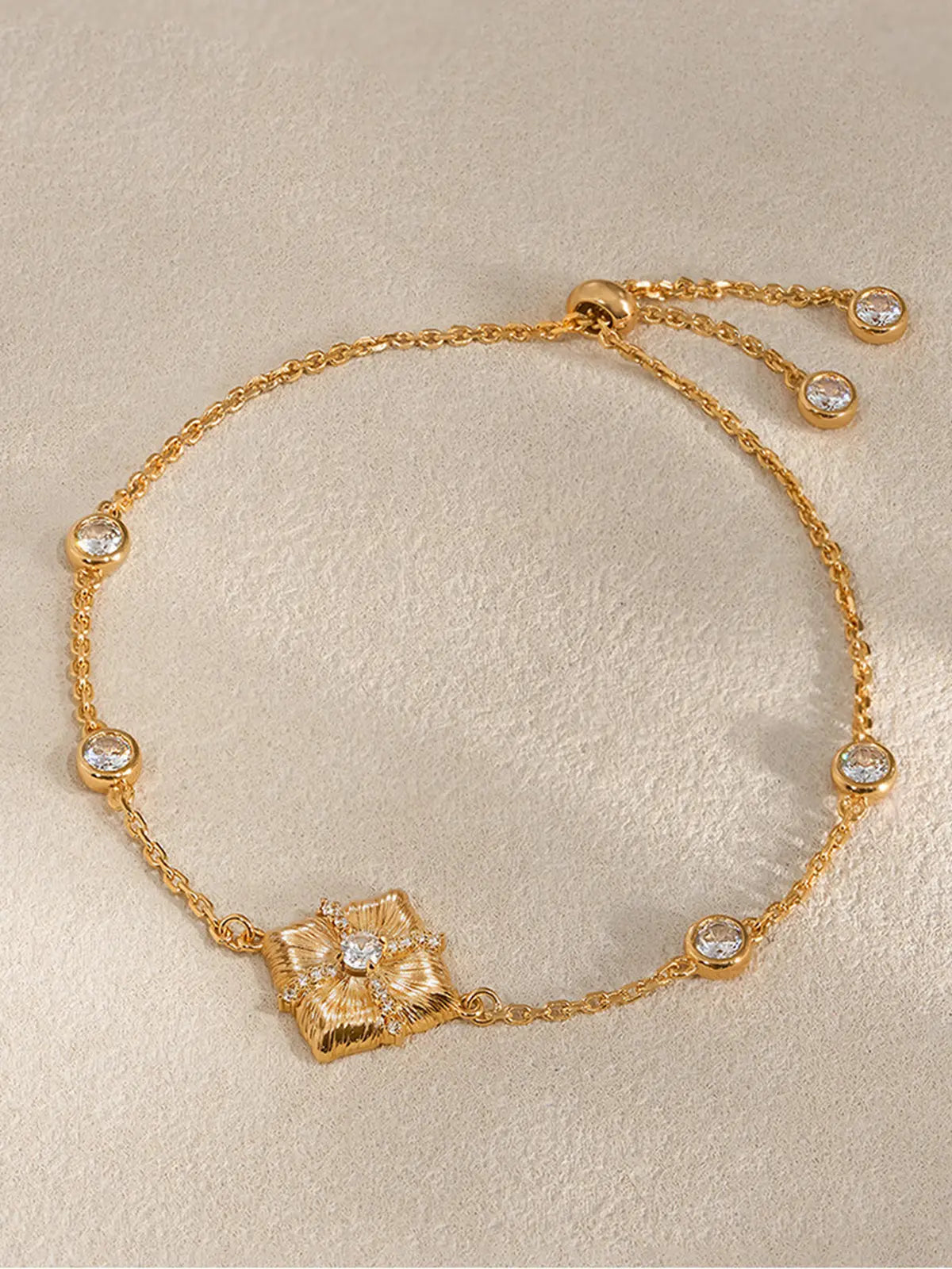 Tactilery Sugar Cube Bracelet in polished gold displayed lying flat on a warm cream textured surface, the full adjustable slider bracelet shown face-on with the large square diamond-shaped center pendant with four deeply ribbed radiating petal-like lobes, central prong-set large round zircon, and pavé zircon cross accent, on a fine polished gold cable chain with four bezel-set round zircon stations and the gold bead slider with two bezel-set round zircon charm ends visible
