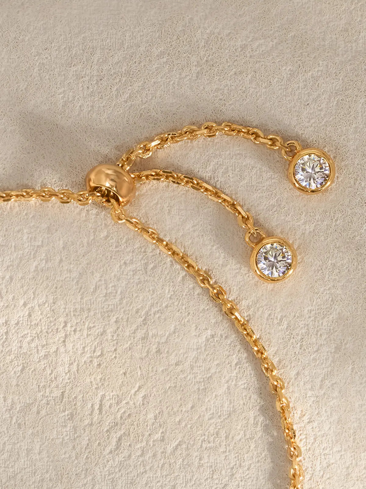 Extreme close-up of the Tactilery Sugar Cube Bracelet adjustable closure on a warm cream textured surface, showing the polished gold ball bead slider mechanism with two extending gold cable chain tails each terminating in a bezel-set round zircon charm