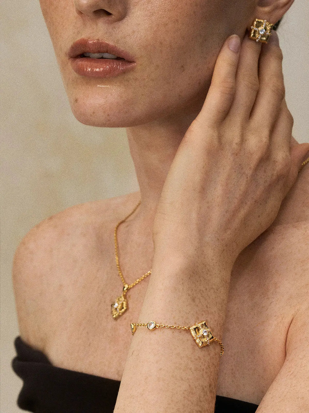 Three-quarter portrait of a model wearing the Tactilery Sugar Cube Bracelet on the wrist with hand raised to cheek, the large square diamond-shaped gold pendant with four deeply ribbed radiating petal-like lobes and pavé zircon cross accent visible at the wrist, paired with a matching sugar cube pendant necklace and sugar cube ring, against a warm taupe background wearing a dark strapless top with short light hair