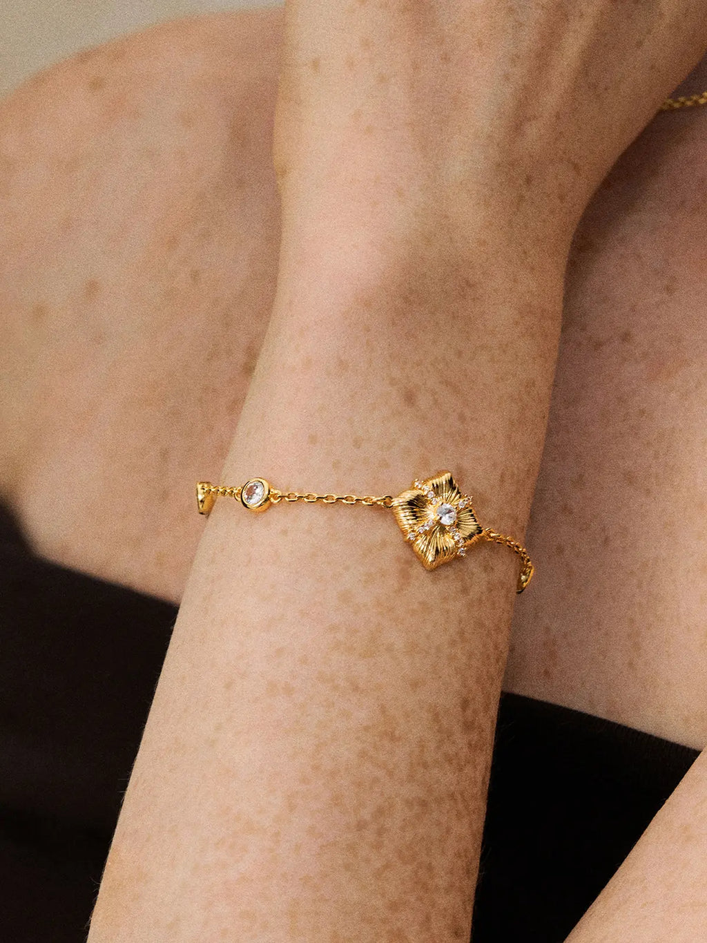 Extreme close-up of a model's wrist wearing the Tactilery Sugar Cube Bracelet, the large square diamond-shaped gold pendant with four deeply ribbed radiating petal-like lobes, central prong-set large round zircon, and pavé zircon cross accent sitting at the wrist on a fine polished gold cable chain with bezel-set round zircon stations, against a warm skin-tone background