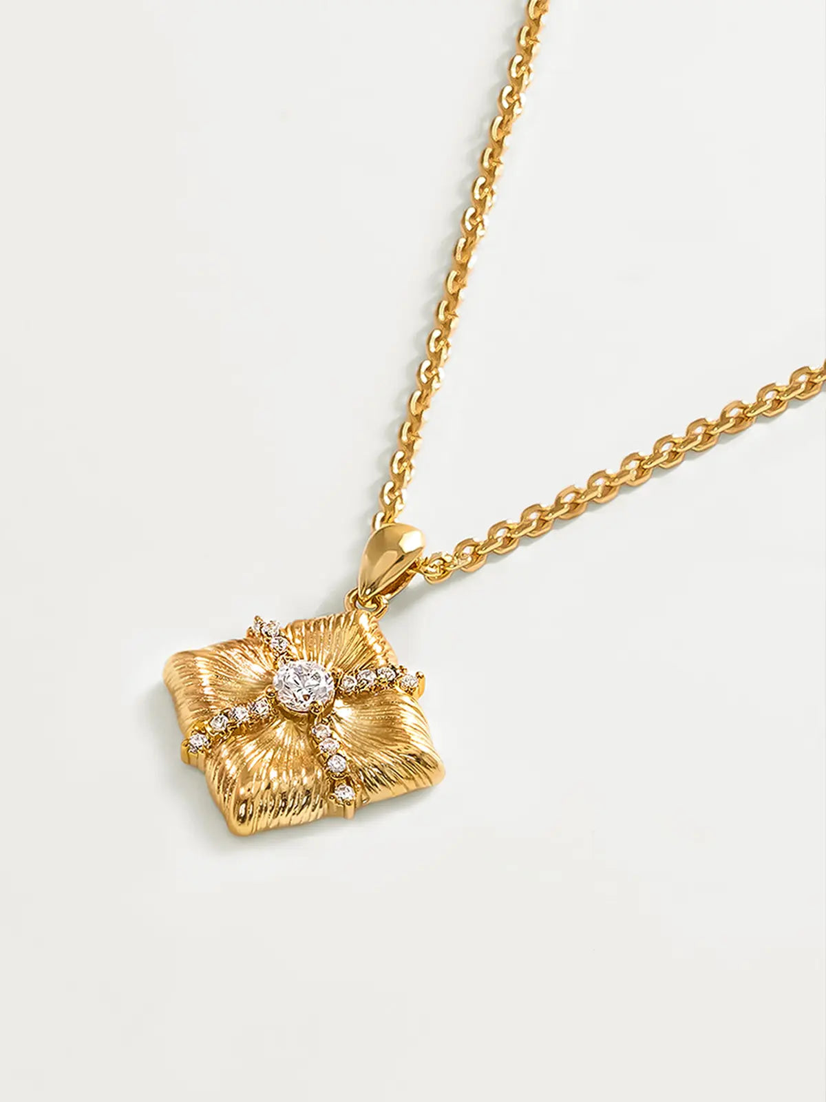 Extreme close-up of the Tactilery Sugar Cube Necklace pendant on a warm cream surface, the square diamond-shaped polished gold pendant with four deeply radiating sunburst line-engraved petal-like lobes filling the frame — the central prong-set large round zircon and pavé zircon cross accent in sharp detail — with the fine polished gold curb chain and bail visible above