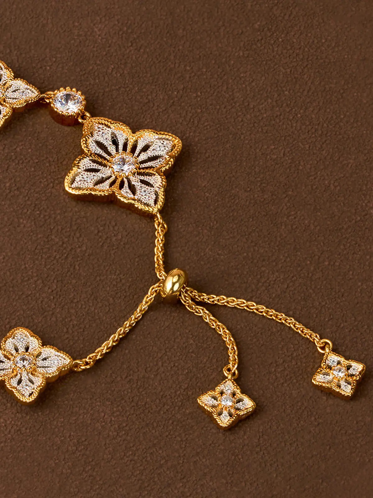 Extreme close-up of the Tactilery Two-Tone Persimmon Calyx Bracelet on a dark brown suede surface, showing the polished gold adjustable bead slider mechanism with the two small two-tone persimmon calyx charm ends on the extending gold rope chain tails, and two large two-tone persimmon calyx flower links with matte brushed silver openwork petal bodies and central prong-set round zircons flanking the slider