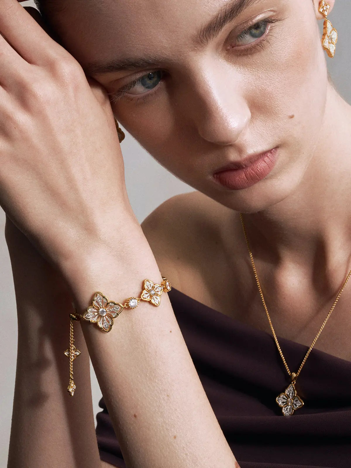 Three-quarter portrait of a model wearing the Tactilery Two-Tone Persimmon Calyx Bracelet on the wrist with hand raised to face and gaze downward, the adjustable polished gold and matte brushed silver persimmon calyx flower link bracelet with central prong-set round zircon accents visible at the wrist, paired with matching two-tone persimmon calyx drop earrings and a gold persimmon calyx pendant necklace, against a warm grey background wearing a dark draped top with dark hair slicked back