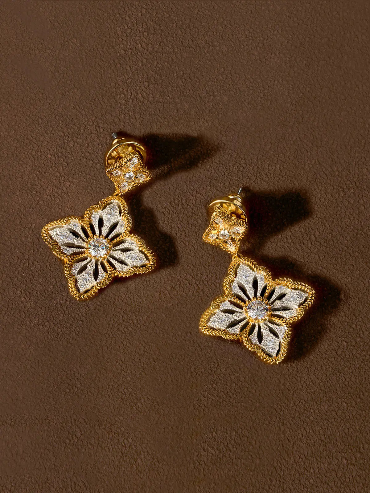 Pair of Tactilery Two-Tone Persimmon Calyx Earrings displayed at an angle on a dark brown suede surface, one showing the full front face of the large four-petal flower drop with matte brushed silver sand-textured petals, rope-twist gold border, and central prong-set round zircon, one showing the three-dimensional depth of the flower form, with silver post pins visible at top